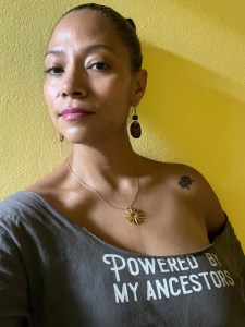 Photo of author Zayda Rivera standing in front of a yellow wall, looking at the camera, wearing a shirt that says “Powered By My Ancestors”